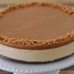 A creamy caramel cheesecake sits on a white fluted cake stand. The cheesecake has a smooth caramel topping and a crumbly crust around the outer edge. The background is a wooden surface.