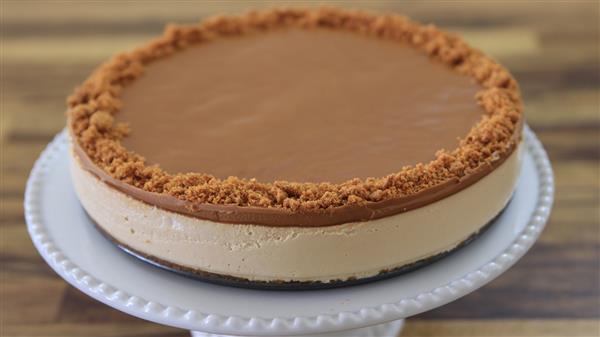 A creamy caramel cheesecake sits on a white fluted cake stand. The cheesecake has a smooth caramel topping and a crumbly crust around the outer edge. The background is a wooden surface.