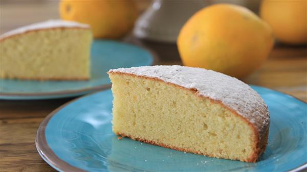 A slice of sponge cake dusted with powdered sugar is placed on a blue plate. In the background, another plate with a slice of cake and some oranges are visible. The scene is set on a wooden surface.