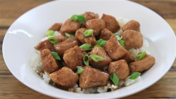 A white plate holds a serving of stir-fried chicken pieces glazed in a brown sauce, garnished with green onion slices. The chicken rests on a bed of white rice, and the background features a wooden surface.