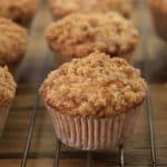 Close-up image of multiple freshly baked muffins cooling on a wire rack. The muffins have a crumbly streusel topping and the golden-brown texture suggests they are moist and well-baked. They are arranged in even rows, highlighting their uniform size and shape.