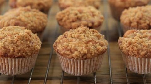 Close-up image of multiple freshly baked muffins cooling on a wire rack. The muffins have a crumbly streusel topping and the golden-brown texture suggests they are moist and well-baked. They are arranged in even rows, highlighting their uniform size and shape.