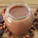 A glass jar filled with creamy chocolate spread is placed on a wooden surface. Surrounding the jar are whole hazelnuts and pieces of dark chocolate, highlighting the ingredients used in the spread.
