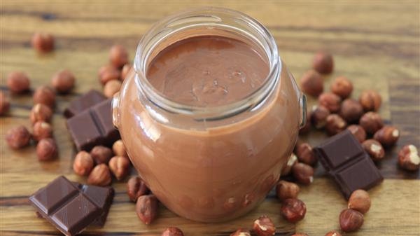 A glass jar filled with creamy chocolate spread is placed on a wooden surface. Surrounding the jar are whole hazelnuts and pieces of dark chocolate, highlighting the ingredients used in the spread.