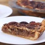 A close-up of a slice of pecan pie on a white plate, placed on a wooden table. The pie has a golden-brown flaky crust with a generous filling of caramelized pecans. Another plate with a slice and a glass pie dish with the rest of the pie are in the background.