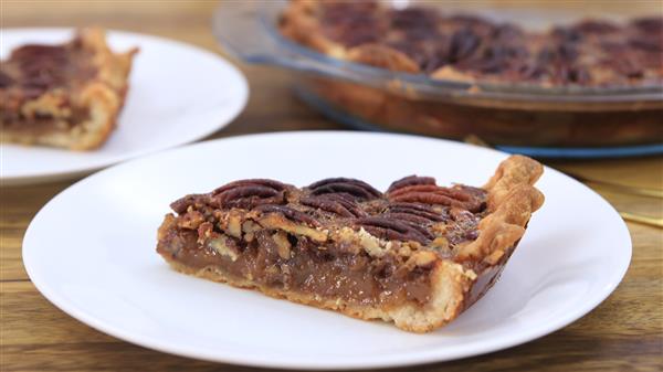 A close-up of a slice of pecan pie on a white plate, placed on a wooden table. The pie has a golden-brown flaky crust with a generous filling of caramelized pecans. Another plate with a slice and a glass pie dish with the rest of the pie are in the background.