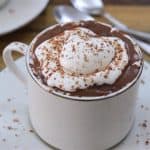 A white ceramic mug filled with hot chocolate, topped with a swirl of whipped cream and sprinkled with chocolate shavings. The mug sits on a matching saucer with chocolate shavings around it. In the background, there is another saucer with two spoons.