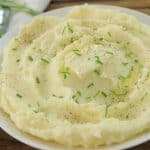 A white plate filled with creamy mashed potatoes garnished with chopped chives and a sprinkle of black pepper, served on a wooden table with a fork and napkin beside it. Fresh green chives are visible in the background.