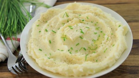 A white plate filled with creamy mashed potatoes garnished with chopped chives and a sprinkle of black pepper, served on a wooden table with a fork and napkin beside it. Fresh green chives are visible in the background.