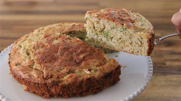 A person lifts a slice of a savory herb and cheese bread from a whole round loaf resting on a white plate. The bread has a golden-brown crust and is speckled with green herbs throughout. The background features a wooden surface.