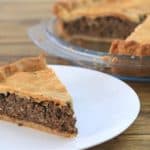 A slice of meat pie on a white plate. In the background, the rest of the pie with a golden-brown crust sits in a clear glass pie dish on a wooden surface. The meat filling is visible, and the crust appears flaky and well-baked.