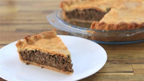 A slice of meat pie on a white plate. In the background, the rest of the pie with a golden-brown crust sits in a clear glass pie dish on a wooden surface. The meat filling is visible, and the crust appears flaky and well-baked.