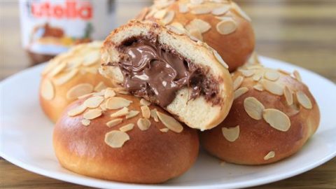 A white plate holds several round buns topped with almond slices. The front-center bun is cut open, revealing a rich chocolate hazelnut filling that is slightly oozing out. A Nutella jar is blurred in the background.