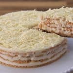 A layered cake with visible cream and sponge cake layers is topped with white chocolate shavings. A slice is being lifted from the cake with a metal spatula. The cake sits on a white cake stand, with a wooden surface in the background.