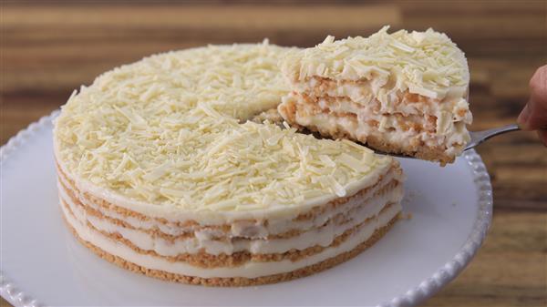 A layered cake with visible cream and sponge cake layers is topped with white chocolate shavings. A slice is being lifted from the cake with a metal spatula. The cake sits on a white cake stand, with a wooden surface in the background.