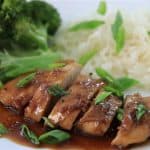 A white plate with a serving of sliced teriyaki chicken topped with green onions, steamed white rice, and broccoli florets. The teriyaki sauce glistens over the chicken, and the dish is presented neatly with vibrant colors.