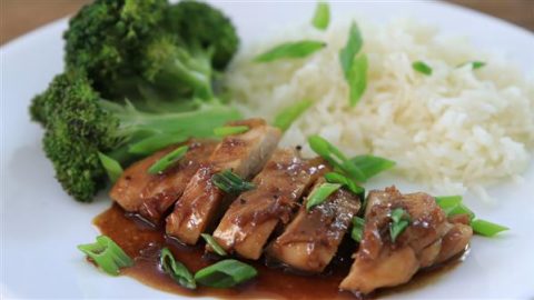 A white plate with a serving of sliced teriyaki chicken topped with green onions, steamed white rice, and broccoli florets. The teriyaki sauce glistens over the chicken, and the dish is presented neatly with vibrant colors.