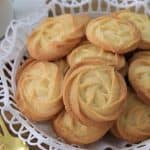 A basket filled with golden, swirl-patterned butter cookies is placed on a wooden table. The basket is lined with a white doily. In the background, there is a white teacup and saucer, and a couple of ornate gold spoons are resting beside the basket.