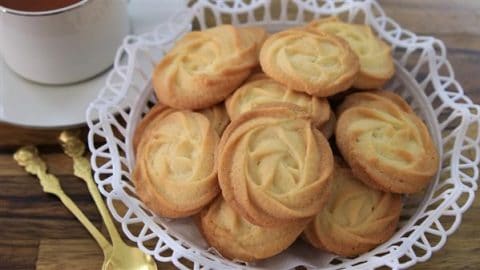 A basket filled with golden, swirl-patterned butter cookies is placed on a wooden table. The basket is lined with a white doily. In the background, there is a white teacup and saucer, and a couple of ornate gold spoons are resting beside the basket.