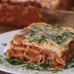 A close-up of a slice of lasagna on a white plate. The lasagna has visible layers of pasta, meat sauce, and cheese, topped with finely chopped parsley and grated Parmesan. In the background, a baking dish with the remaining lasagna is partially visible.