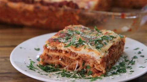 A close-up of a slice of lasagna on a white plate. The lasagna has visible layers of pasta, meat sauce, and cheese, topped with finely chopped parsley and grated Parmesan. In the background, a baking dish with the remaining lasagna is partially visible.