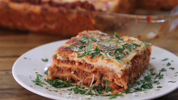 A close-up of a slice of lasagna on a white plate. The lasagna has visible layers of pasta, meat sauce, and cheese, topped with finely chopped parsley and grated Parmesan. In the background, a baking dish with the remaining lasagna is partially visible.