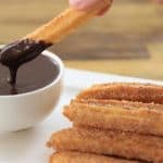 A close-up of several sugar-coated churros stacked on a white plate. A hand is dipping one churro into a small white bowl of thick chocolate sauce. The background is blurred, showing a wooden surface.