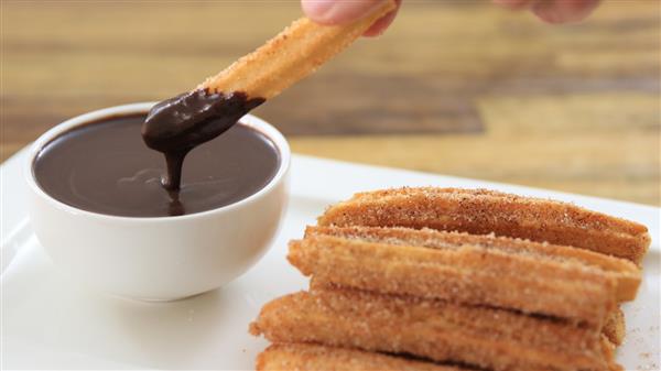 A close-up of several sugar-coated churros stacked on a white plate. A hand is dipping one churro into a small white bowl of thick chocolate sauce. The background is blurred, showing a wooden surface.