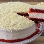 A red velvet cake with cream cheese frosting and white chocolate shavings. One slice has been cut and is being lifted, displaying the red and cream layers inside. The cake is on a decorative white plate with scalloped edges.