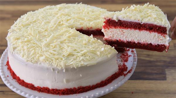 A red velvet cake with cream cheese frosting and white chocolate shavings. One slice has been cut and is being lifted, displaying the red and cream layers inside. The cake is on a decorative white plate with scalloped edges.