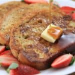 A plate of French toast topped with a pat of butter and drizzled with syrup. Fresh strawberry slices are arranged around the toast on the white plate, adding a touch of color to the dish. The French toast appears golden brown and inviting.