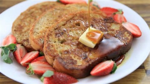 A plate of French toast topped with a pat of butter and drizzled with syrup. Fresh strawberry slices are arranged around the toast on the white plate, adding a touch of color to the dish. The French toast appears golden brown and inviting.