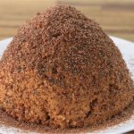 A white plate holds a round, dome-shaped dessert covered in finely grated chocolate. The dessert has a crumbly texture and is placed on a wooden table, with some of the grated chocolate scattered around the plate.