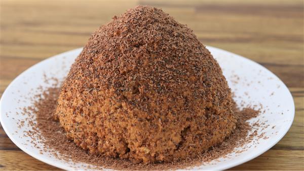 A white plate holds a round, dome-shaped dessert covered in finely grated chocolate. The dessert has a crumbly texture and is placed on a wooden table, with some of the grated chocolate scattered around the plate.