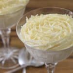 Two elegant glass dessert cups filled with creamy white mousse, topped with shavings of white chocolate. The cups are set on a wooden surface, and a spoon is placed between them.