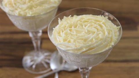 Two elegant glass dessert cups filled with creamy white mousse, topped with shavings of white chocolate. The cups are set on a wooden surface, and a spoon is placed between them.