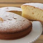 A round, golden-brown cake dusted with powdered sugar sits on a white cake stand. A slice has been cut and is being lifted out, revealing a soft, fluffy, light-yellow interior. The background features a wooden surface.