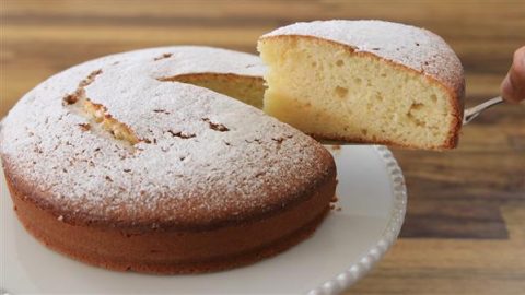 A round, golden-brown cake dusted with powdered sugar sits on a white cake stand. A slice has been cut and is being lifted out, revealing a soft, fluffy, light-yellow interior. The background features a wooden surface.