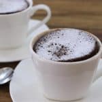 Two white cups are filled with chocolate soufflé and dusted with powdered sugar, placed on white saucers with metal teaspoons beside them. The setting is on a wooden table, giving a cozy and inviting feel.