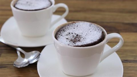 Two white cups are filled with chocolate soufflé and dusted with powdered sugar, placed on white saucers with metal teaspoons beside them. The setting is on a wooden table, giving a cozy and inviting feel.