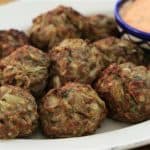 A plate filled with several crispy, golden-brown meatballs is shown. A small bowl of creamy dipping sauce with a pinkish hue sits on the edge of the plate. The meatballs appear to have herbs and other ingredients mixed in.