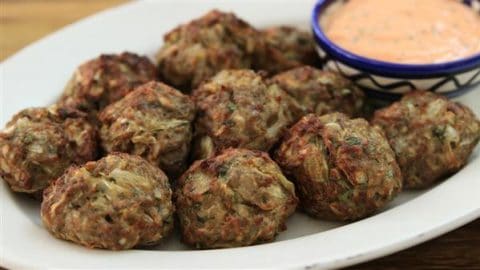 A plate filled with several crispy, golden-brown meatballs is shown. A small bowl of creamy dipping sauce with a pinkish hue sits on the edge of the plate. The meatballs appear to have herbs and other ingredients mixed in.