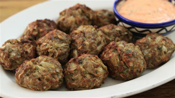 A plate filled with several crispy, golden-brown meatballs is shown. A small bowl of creamy dipping sauce with a pinkish hue sits on the edge of the plate. The meatballs appear to have herbs and other ingredients mixed in.