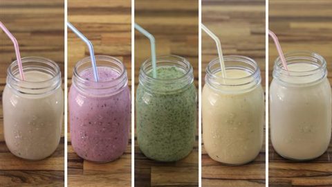 Five mason jars filled with different fruit smoothies, each with a straw. From left to right: a light beige smoothie, a purple smoothie, a green smoothie, a pale yellow smoothie, and a yellow smoothie. They are lined up on a wooden surface.