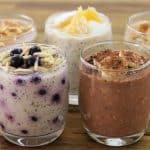 Five glasses of overnight oats are displayed on a wooden surface. Each glass contains a different variety, topped with ingredients such as blueberries, nuts, fruit slices, and cocoa powder. The oats vary in color and texture, indicating different flavors.