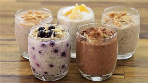 Five glasses of overnight oats are displayed on a wooden surface. Each glass contains a different variety, topped with ingredients such as blueberries, nuts, fruit slices, and cocoa powder. The oats vary in color and texture, indicating different flavors.