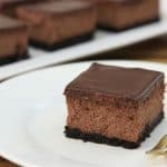 A slice of chocolate cheesecake with a chocolate ganache topping and a cookie crust sits on a white plate with a gold fork beside it. Additional slices are visible on a white platter in the background. The surface is wooden.