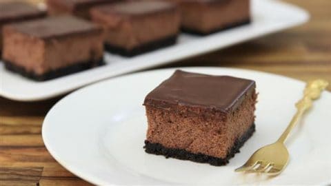 A slice of chocolate cheesecake with a chocolate ganache topping and a cookie crust sits on a white plate with a gold fork beside it. Additional slices are visible on a white platter in the background. The surface is wooden.