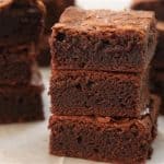 A stack of rich, fudgy chocolate brownies on a light-colored surface. The brownies are freshly cut into square pieces, showcasing their moist and dense texture. More brownie squares are visible in the background.