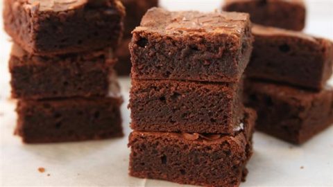 A stack of rich, fudgy chocolate brownies on a light-colored surface. The brownies are freshly cut into square pieces, showcasing their moist and dense texture. More brownie squares are visible in the background.